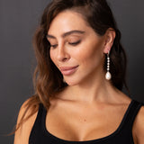 A woman wearing a long, dangling pearl earring with small silver-toned spacer beads, shown in a close-up portrait against a dark gray background.