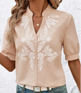 A woman wearing a beige short-sleeve button-front blouse with white floral and leaf embroidery, paired with blue jeans and a white belt, against a textured white wall.