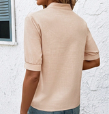 A woman shown from the back wearing a light beige short-sleeve linen shirt with rolled cuffs, paired with blue jeans, standing against a white textured wall.