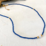 A delicate beaded necklace featuring small blue faceted beads, accented with tiny gold spacers and a single white pearl bead, displayed on a textured off-white surface.