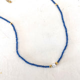 A delicate beaded necklace featuring small faceted blue beads, accented with tiny gold spacers and a single white pearl bead, displayed on a textured off-white surface.