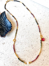 A delicate beaded necklace with gold-toned chain links, alternating beads of dark green, pink, and forrest, featuring a small gold heart charm and a round pink stone accent, displayed on a light textured surface next to a dark wing-shaped decorative object.