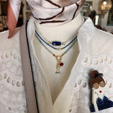 A close-up of a layered necklace set displayed on a cream mannequin torso. The set includes a pearl choker, a blue beaded double-strand chain with a central oval blue stone and gold-toned links, a light blue beaded chain, and a gold-toned pendant necklace with a fish charm and a small red bead.
