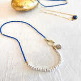 A beaded necklace with a vibrant blue top section, a strand of white shell beads, and a gold clasp with a small gray stone charm, displayed on a textured off-white surface next to a gold leaf ornament.