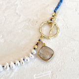 A close-up of a beaded necklace featuring white baroque pearls, blue faceted beads, a gold toggle clasp, and a square druzy stone pendant on a textured off-white background.
