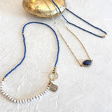 Three delicate beaded necklaces arranged on a textured white surface with a gold stone prop. One necklace has blue beads and a silver-toned toggle clasp with a gray stone pendant.