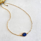 A delicate gold chain necklace featuring an oval blue gemstone pendant with a small clear stone border, displayed on a textured white surface.