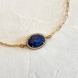 A gold chain necklace featuring an oval blue lapis lazuli pendant with a halo of small clear stones, displayed on a textured off-white background.