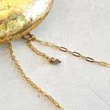A close-up of a gold chain necklace with a paperclip-style extender and a small gray bead, laid on a light textured surface with part of a gold decorative object visible in the upper left.