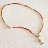 A beaded necklace featuring small graded orange faceted beads, accented with a single white pearl and a gold-tone fish charm, displayed on a textured off-white surface.