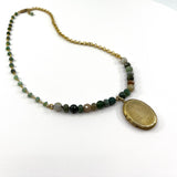 A vintage-style beaded necklace featuring a gold chain, a row of green and earthy stone beads, and a large oval gold locket pendant, displayed on a clean white background.