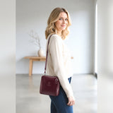A woman wearing a cream knit sweater and blue jeans carries a burgundy leather crossbody bag with a matching strap and a small gold logo on the front, set in a bright, modern minimalist room.