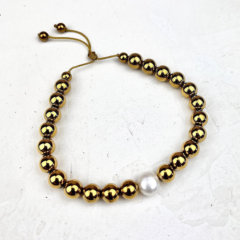 Gold beaded bracelet with a single white pearl on a light gray background