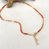 A beaded necklace featuring graded orange carnelian beads, a small white pearl, gold-toned spacer beads, and a gold-toned fish charm pendant, displayed on a textured off-white surface.