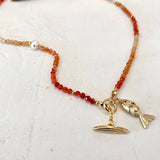 A delicate beaded necklace with small orange-red and cream beads, featuring a gold-tone fish charm and a small gold-tone boat charm, displayed on a light textured surface.