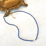 A delicate beaded necklace with deep blue beads, small gold spacer beads, and a single white pearl accent, displayed on a textured white surface next to an ornate gold tray.