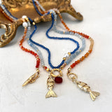 Three beaded necklaces with gold fish charms and a central gold clasp, displayed on a white surface with a decorative gold tray in the background. The necklaces feature red, light blue, and dark blue beads.