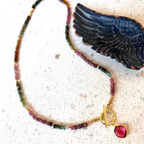 A beaded necklace with small multicolored gemstone beads, gold-toned spacer accents, a gold toggle clasp, and a round pink gemstone pendant, displayed on a light stone surface next to a black wing-shaped object.