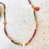 A delicate beaded necklace with gold-toned beads, small green and pink gemstone beads, a white pearl-like bead, and a pink oval stone charm, displayed on a light textured surface.