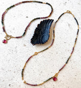 A beaded necklace with gold-toned links, multicolored beads, and small pink gemstone charms, displayed on a light textured surface alongside a dark carved wing-shaped pendant.