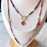 A layered beaded necklace displayed on a cream fabric jewelry bust. The necklace features small pink, green, and gold beads, a gold toggle clasp, and a dangling pink stone pendant.