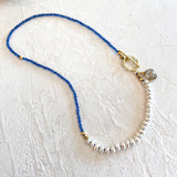 A beaded necklace featuring a vibrant blue top section, a transition of small white beads, a gold-toned clasp, and a gray faceted stone charm, displayed on a textured off-white background.