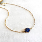 A delicate gold chain necklace featuring an oval blue gemstone pendant with a small border of clear stones, displayed on a textured white surface.