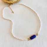 A delicate pearl necklace with a central oval blue gemstone surrounded by small clear stones and gold-tone settings, displayed on a light textured surface.