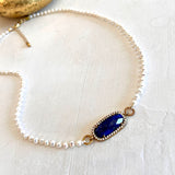 A delicate necklace featuring a strand of small white pearls and a central oval blue gemstone set in gold-toned metal with small clear stones around it, displayed on a textured off-white surface.