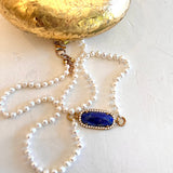 A pearl necklace with a gold-tone oval pendant featuring a deep blue cabochon stone surrounded by small clear crystals, displayed on a textured off-white surface with a gold decorative object at the top.