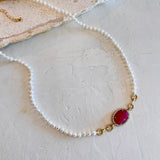 A delicate white pearl necklace with gold chain links and a central oval magenta-pink cabochon stone, displayed on a textured off-white surface.