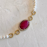 A close-up of a delicate bracelet featuring a central oval pinkish-red gemstone surrounded by small clear stones, set in gold-toned metal with twisted chain links, and a strand of white pearl-like beads on each end, displayed on a textured off-white surface.