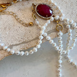 A close-up of a pearl necklace with a gold-tone clasp and extender chain, featuring a large oval dark red gemstone pendant surrounded by small clear stones, displayed on a textured light stone surface.