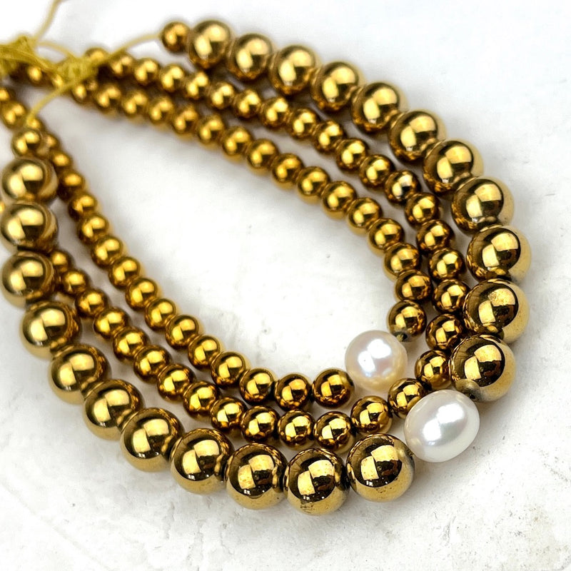 Gold beaded bracelets with white pearls on a textured white background