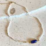 A delicate necklace with small white pearls and a central oval blue stone set in gold, displayed on a textured beige stone surface.