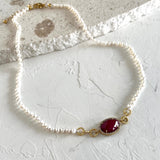 A delicate necklace made of small white pearls with gold-toned links and a central oval red gemstone, photographed on a textured white surface.