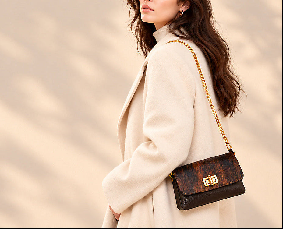 A woman in a cream coat carries a dark brown textured leather shoulder bag with a gold chain strap, set against a warm beige wall with soft leaf shadows.